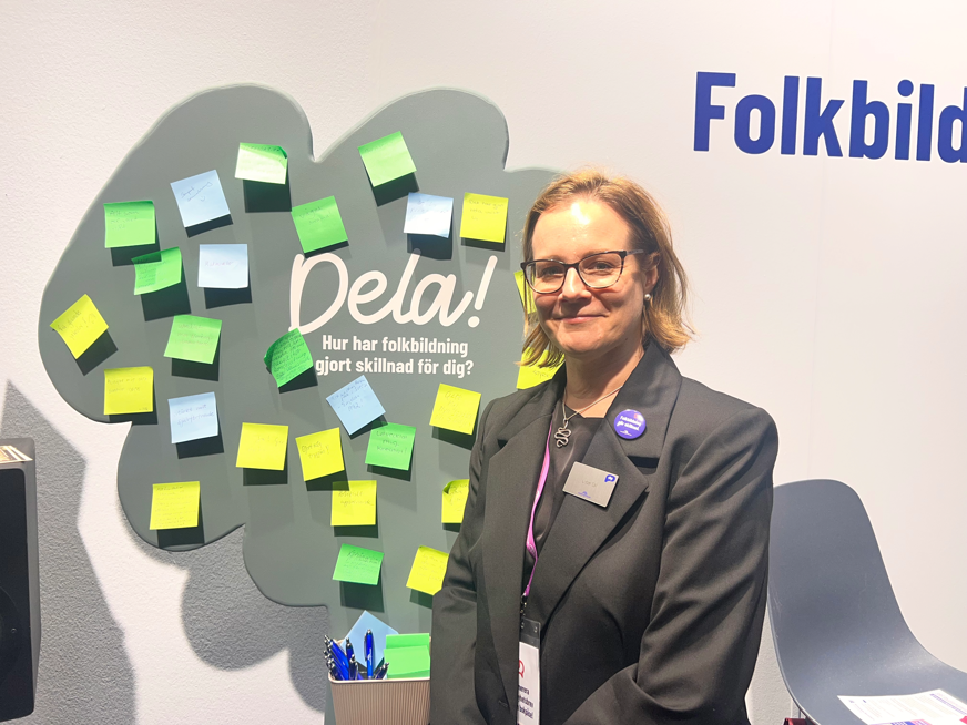 Lisbet Säll står framför en vägg med en stor skylt för folkbildning. På skylten står bland annat ”Dela! Hur har folkbildning gjort skillnad för dig?” och den är täckt av färgglada post-it-lappar. Lisbet bär glasögon och mörk kavaj med en namnbricka och en lila knapp på bröstet. Bredvid henne står en stol och framför skylten finns en behållare med pennor.