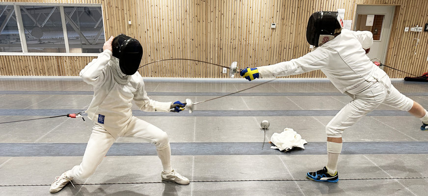 Två fäktare i rörelse. Den vänstra hukar sig ner och riktar värjan mot motståndaren. Den högra gör en framåtrörelse med bakre benet i luften och sticker värjan på bröstet på den vänstra fäktaren.