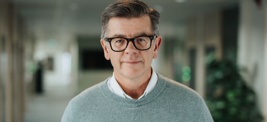 Lars Arrhenius har kort, mörkt hår och svarta, rektangulära glasögon står vänd mot kameran. Han har en ljusgrå stickad tröja över en vit skjorta. Bakgrunden är en ljus, suddig inomhusmiljö med gröna växter.