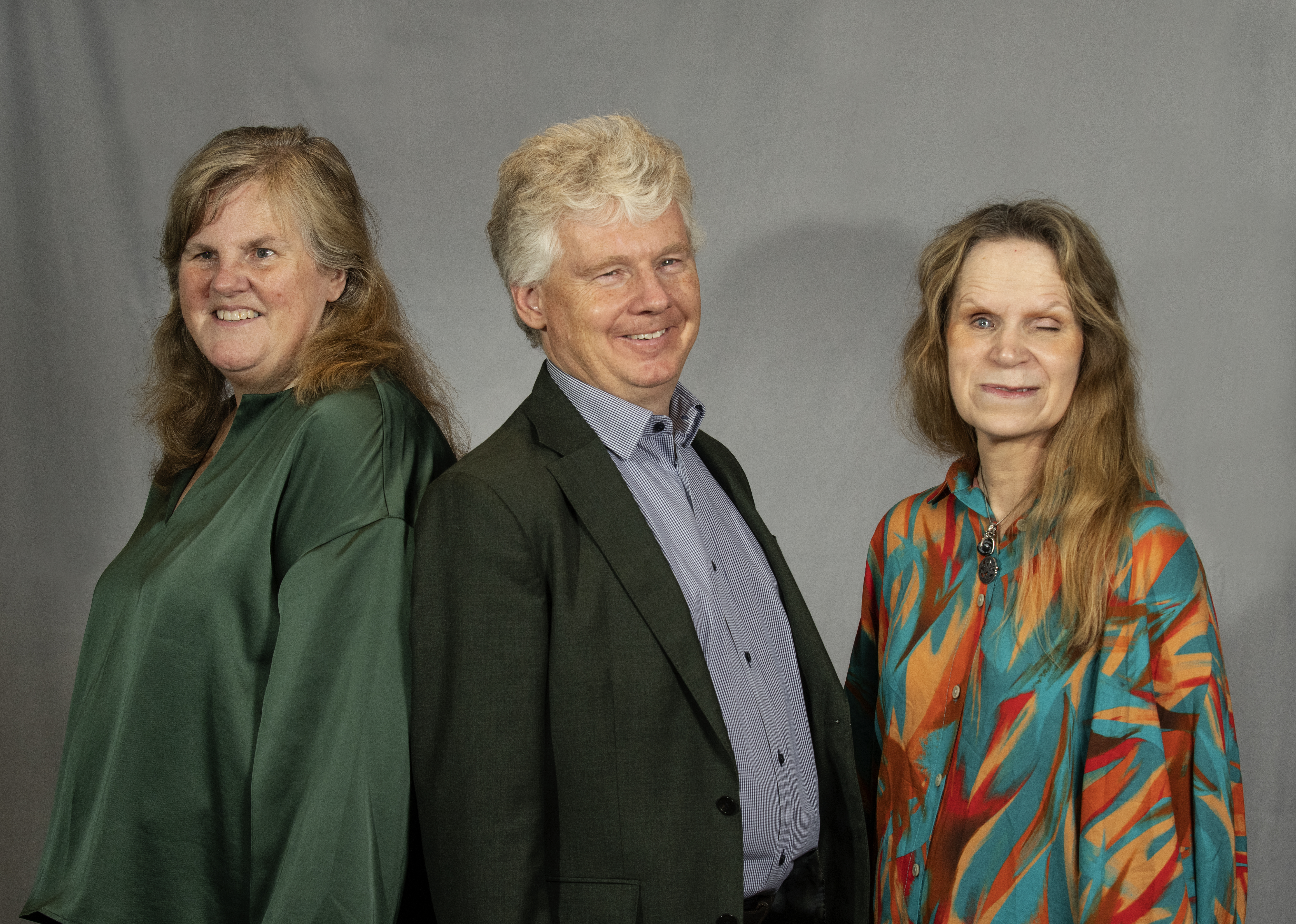 En man och två kvinnor poserar framför en fotovägg. Kvinnorna är färgglatt klädda och mannen har mörk kostym och ljusblå skjorta.