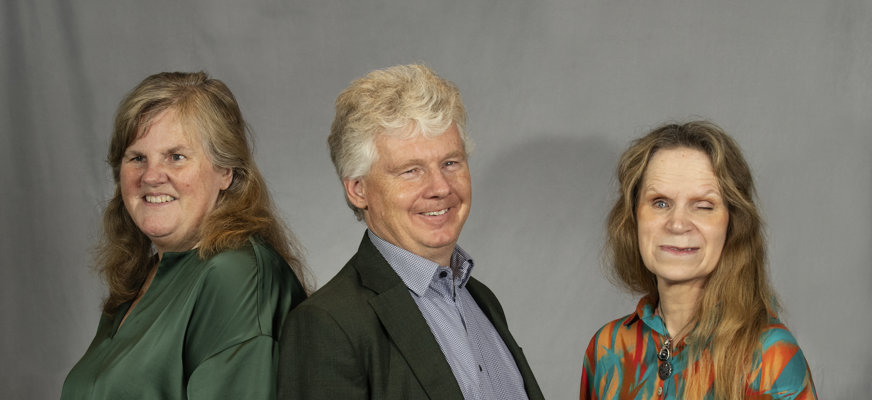 En man och två kvinnor poserar framför en fotovägg. Kvinnorna är färgglatt klädda och mannen har mörk kostym och ljusblå skjorta.