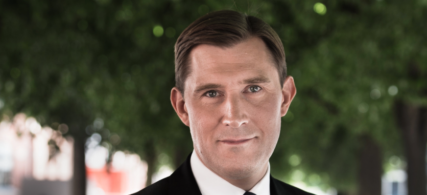 En närbild av Henrik Fritzon i kostym och vit skjorta. Han tittar rakt mot kameran och har kort, bakåtkammat hår och ett neutralt ansiktsuttryck. Bakgrunden är suddig med gröna träd.