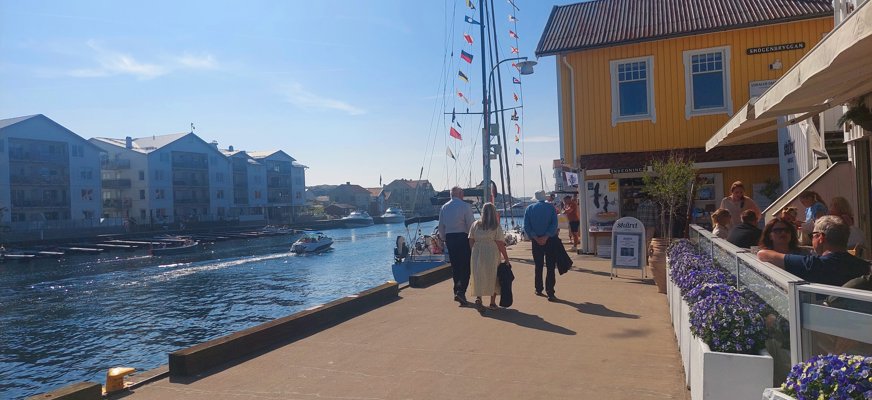 3 personer promenerar på en kaj till vänster en kanal med båtar och på andra sidan hus. Till höger några som sitter på en uteservering, i bakgrunden ett gult hus samt en båtmast med många små flaggor längst masten.