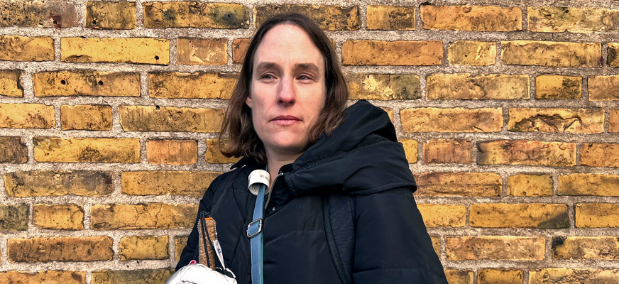 Porträttbild av Eva som står mot en gul tegelvägg. Eva har mörkbrunt hår och mörka ögon Hon är klädd i en svart jacka, reflexhandskar och har en blå väska över axeln. I Evas hand skymtar en vit käpp och hon har ett bekymrat uttryck i ansiktet.