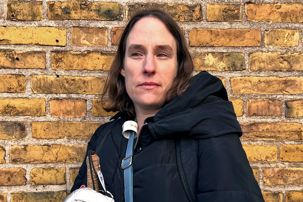 Porträttbild av Eva som står mot en gul tegelvägg. Eva har mörkbrunt hår och mörka ögon Hon är klädd i en svart jacka, reflexhandskar och har en blå väska över axeln. I Evas hand skymtar en vit käpp och hon har ett bekymrat uttryck i ansiktet.