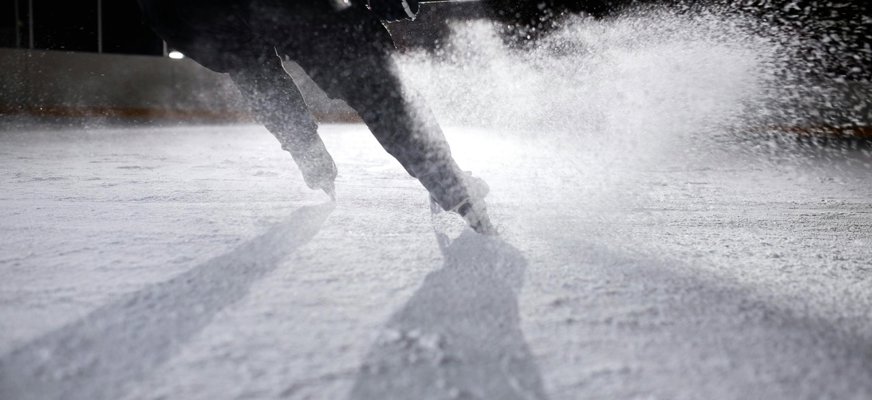 En hockeyspelare klädd i svart syns bakifrån. Han åker skridskor med klubba i handen och skvätter snö som samlats på isen. Bilden är svartvit och tagen i motljus.