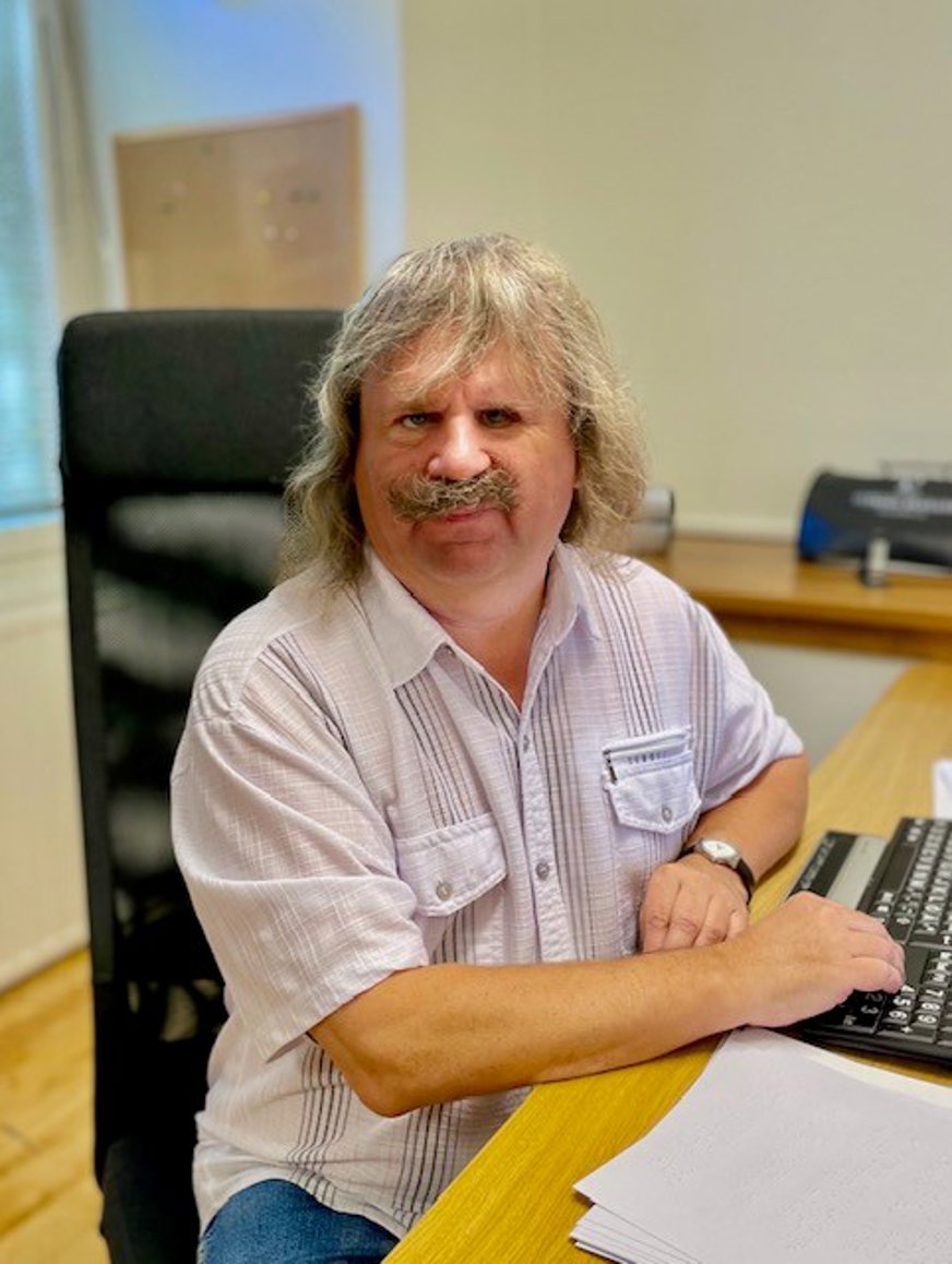 Håkan Thomsson på sitt kontor sitter vid skrivbordet, jeans, vit kortärmad skjorta, grått hår och mustasch