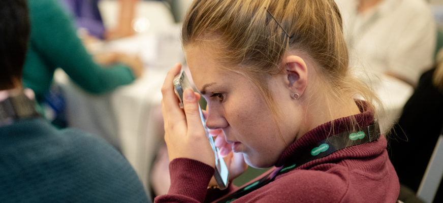 En ung kvinna med blont hår uppsatt i en knut sitter bland andra personer och håller en mobiltelefon mot ansiktet. Hon bär en vinröd tröja och har ett band runt halsen.