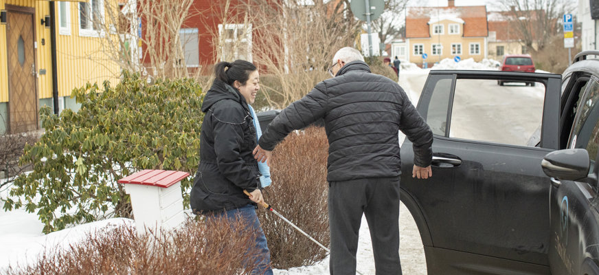 En bild på en kvinna med vit käpp och svart hästsvans som blir hjälpt in i en färdtjänstbild av en chaufför. Chauffören har mörk jacka och grått kort hår.