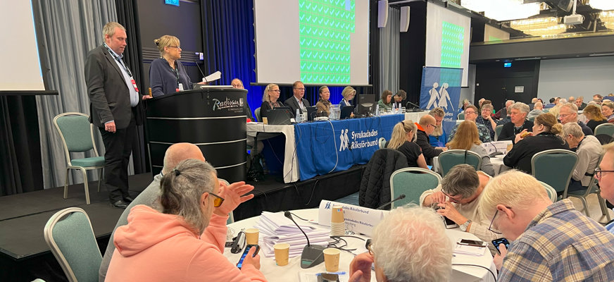 Kongresslokalen med en mängd runda bord där det sitter människor, ett podium där två personer står, samt ett avlångt bord mitt i, där presidiet sitter, försedd en lång blå duk med Synskadades Riksförbunds logotyp.