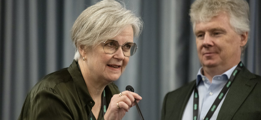 Podium, gråhårig kvinna med tonade glasögon lutar sig mot mikrofonen. I bakgrunden gråhårig man i 50-årsåldern