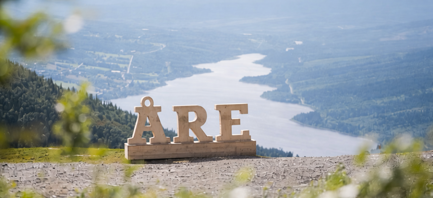 Bilden visar ett vidsträckt fjällandskap i Åre sett från en hög utsiktspunkt. I förgrunden står stora träbokstäver som bildar ordet “ÅRE”, placerade på en träplattform på en grusig yta. Bakom bokstäverna breder landskapet ut sig med Åresjön som slingrar sig genom dalen. Sjön omges av gröna skogar, öppna marker och små bebyggelser som ligger utspridda längs vattnet. Längre bort syns mjuka berg och fjäll som tonar ut i blågrå nyanser i diset.