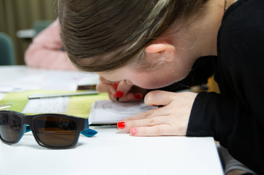 Närbild flicka/ungdom som ritar och lutar sig mycket nära pappret. Ansiktet är vänt bortåt. På bordet papper, solglasögon. Hon har knallrött nagellack.