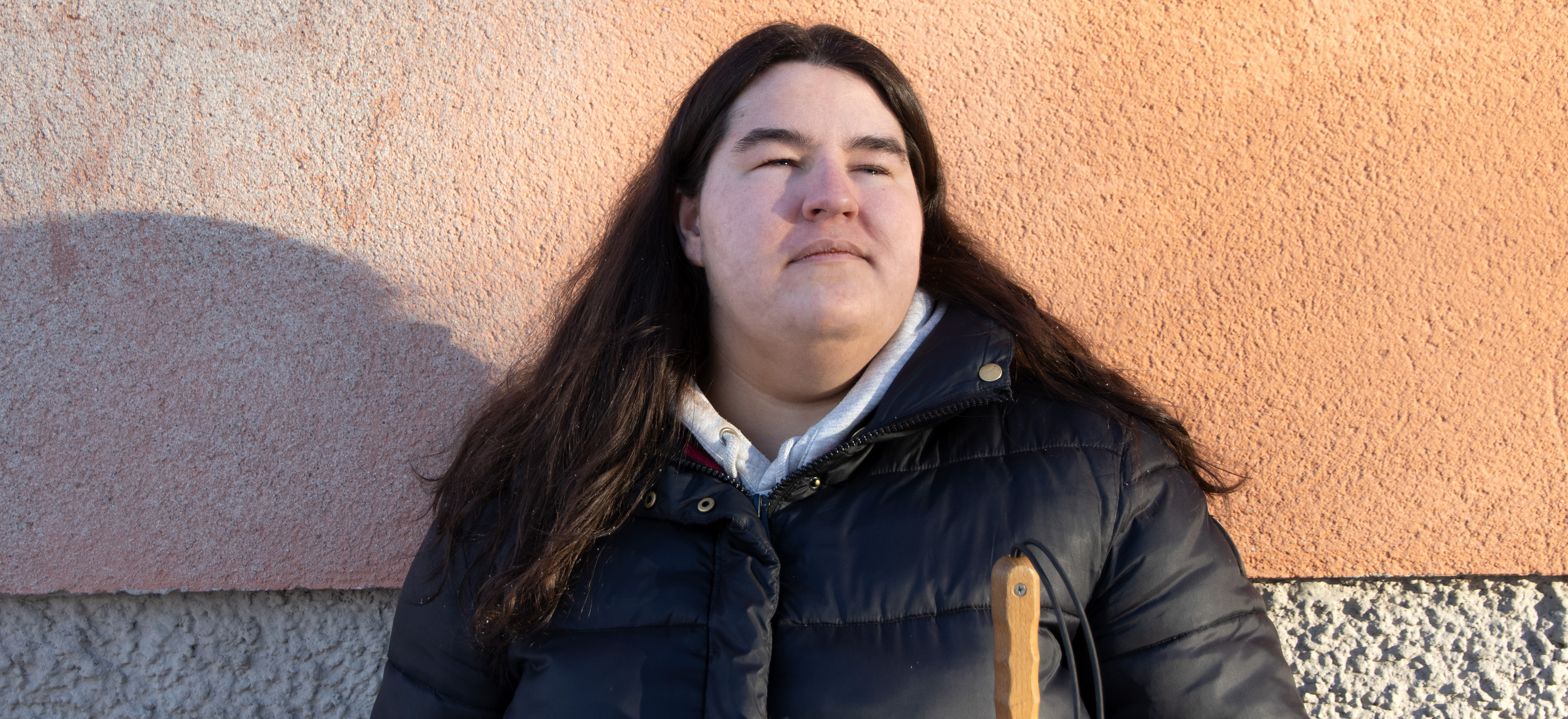 Porträttbild på Oliwia Wisniewska, en ung kvinna i mörkt hår som faller över axlarna. Oliwia står i solen, mot en byggnad med putsad fasad i orange. Hon har en allvarlig uppsyn och blickar uppåt.