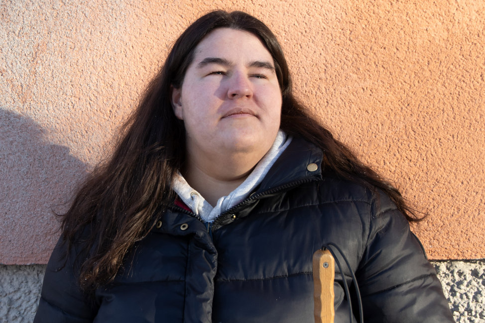 Porträttbild på Oliwia Wisniewska, en ung kvinna i mörkt hår som faller över axlarna. Oliwia står i solen, mot en byggnad med putsad fasad i orange. Hon har en allvarlig uppsyn och blickar uppåt.