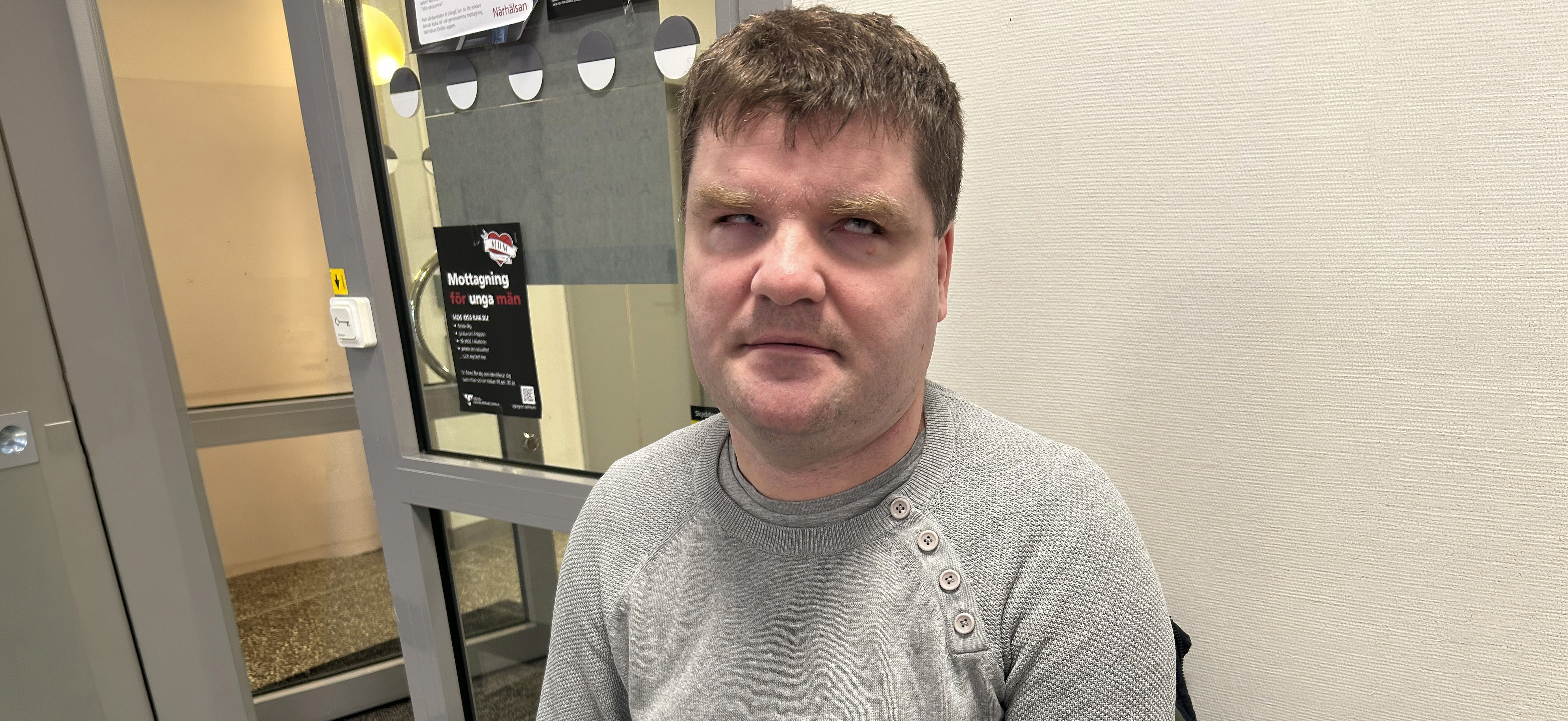 Porträttbild på Damir. Han sitter ute i en korridor på vad som ser ut som en mottagning av något slag. Damir har kort brunt hår, skäggstubb och är klädd i en grå stickad tröja över t-shirt. Det är tydligt på hans ögon att han är synskadad.