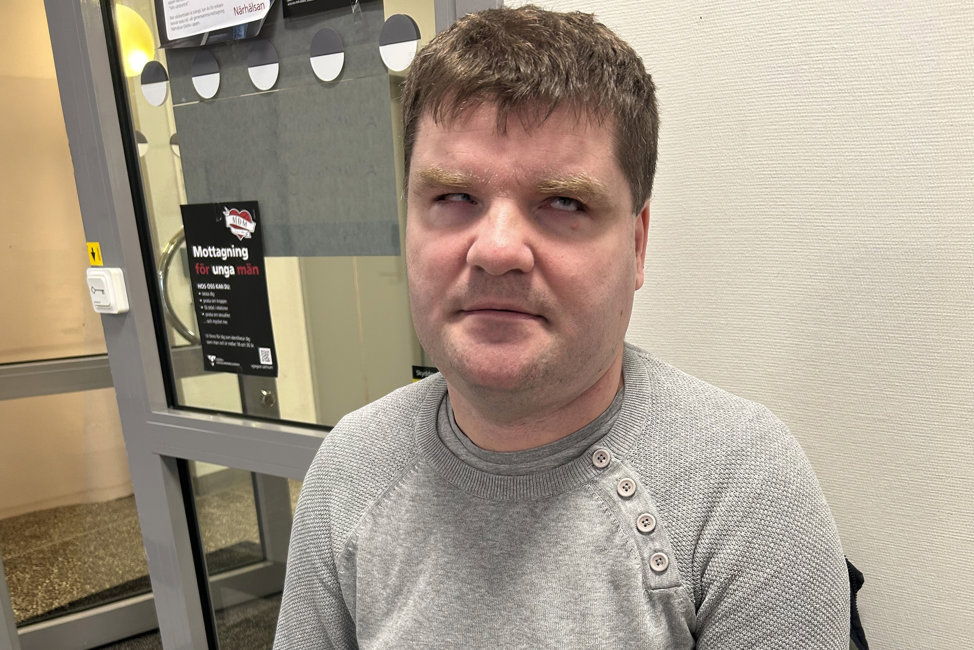 Porträttbild på Damir. Han sitter ute i en korridor på vad som ser ut som en mottagning av något slag. Damir har kort brunt hår, skäggstubb och är klädd i en grå stickad tröja över t-shirt. Det är tydligt på hans ögon att han är synskadad.