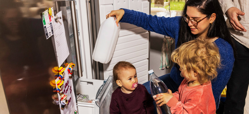 Familj som preppar vattenflaskor och dunkar. De två små barnen hjälper mamman att lägga flaskorna i frysen.