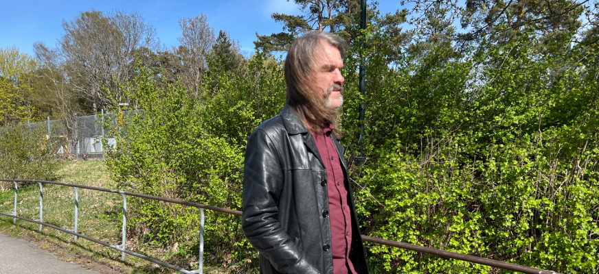 Fredrik, i långt gråbrunt hår och skägg går utomhus i en svart skinnjacka över en vinröd skjorta. Han går framför grönskande buskar och träd i solsken.