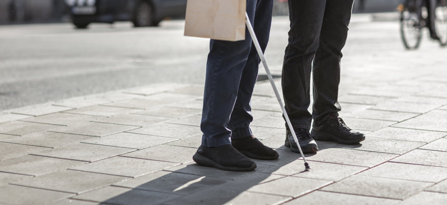 Två personers ben i stadsmiljö, de har båda svarta jeans och står nära varandra. Den ena håller i en vit käpp.
