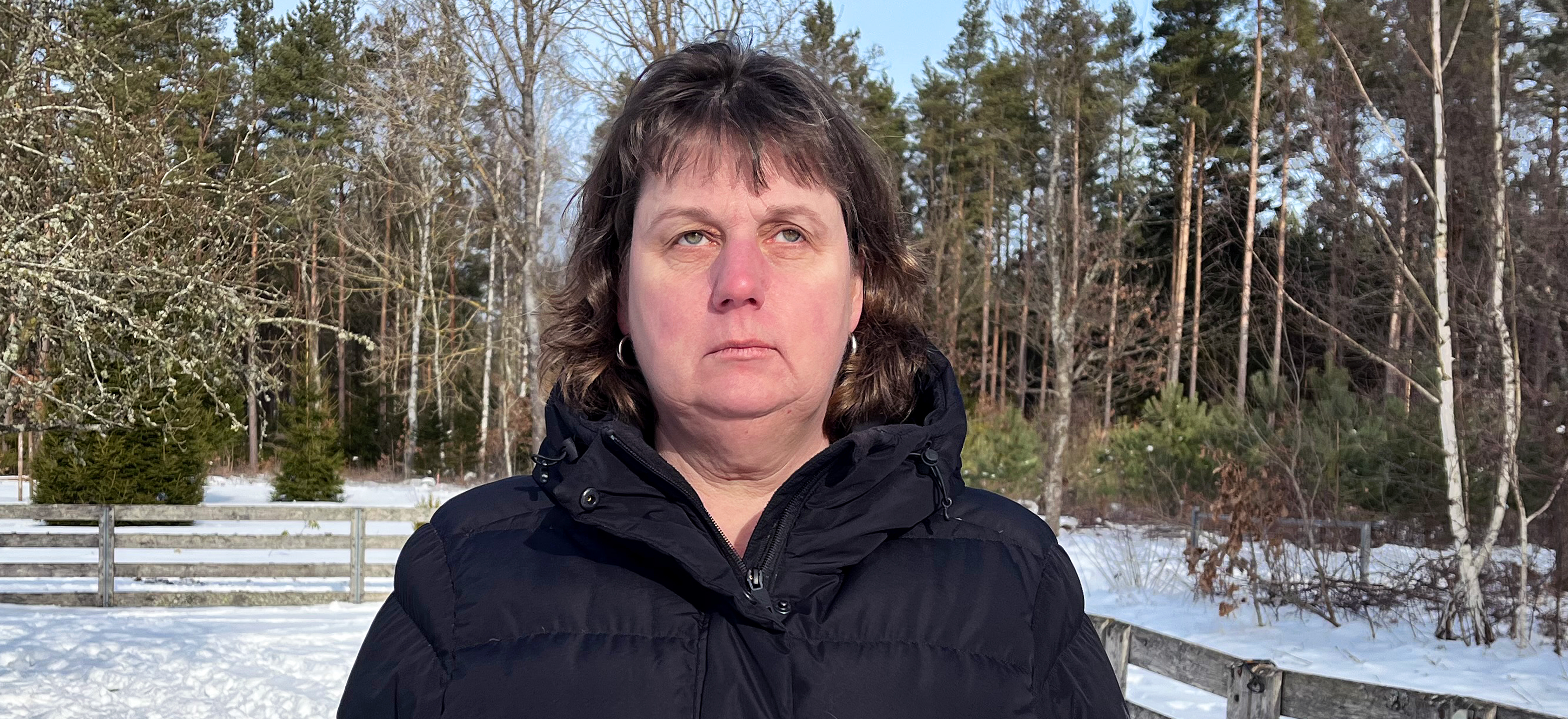 Mari är en medelålders kvinna i brunt, kortklippt hår med lugg. Hon har svart täckjacka och ringar i öronen. Mari står utomhus i en snöig hage och ser med bister min upp mot himlen.