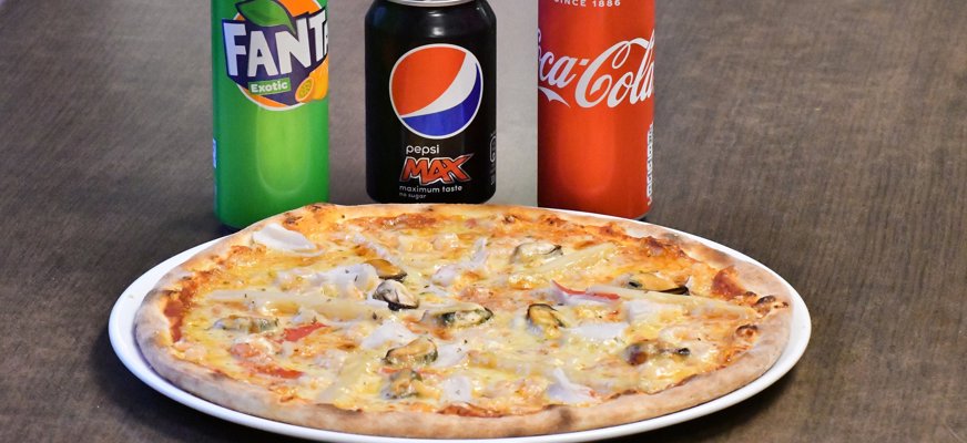 Bilden visar ett mörkt träbord med en pizza på en vit tallrik och 3 läskburkar bakom