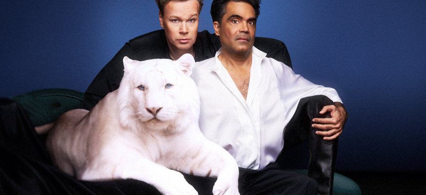 Batra och Glans poserar tillsammans med en vit tiger mot en blå bakgrund. David Batra sitter avslappnat på en pall iklädd en vit skjorta och svarta stövlar, medan Johan Glans står bakom honom med handen på hans axel. Den vita tigern ligger i förgrunden med tassarna vilande över ett svart tygstycke.