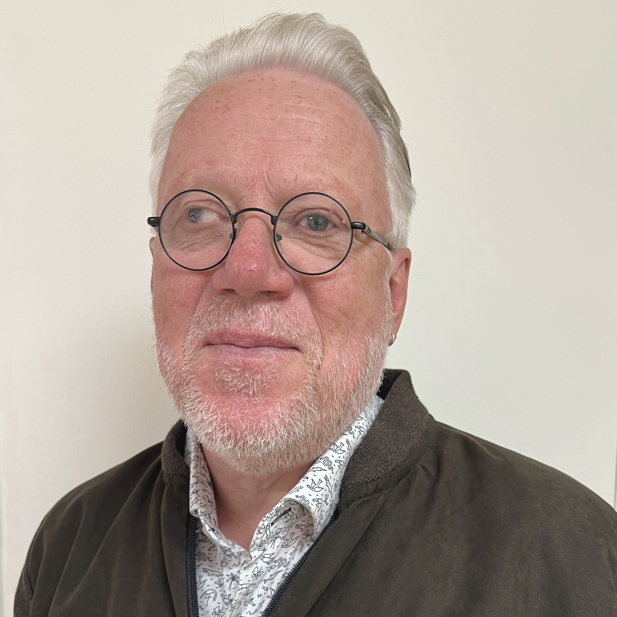Leende man i 60-årsåldern, grått hår, runda glasögon, vitblommig skjorta och kofta.