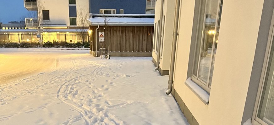 En snöig trottoar i skymningsljus och gatubelysning. Delar av nedersta våningen på två hus och delar av ett cykelförråd syns.