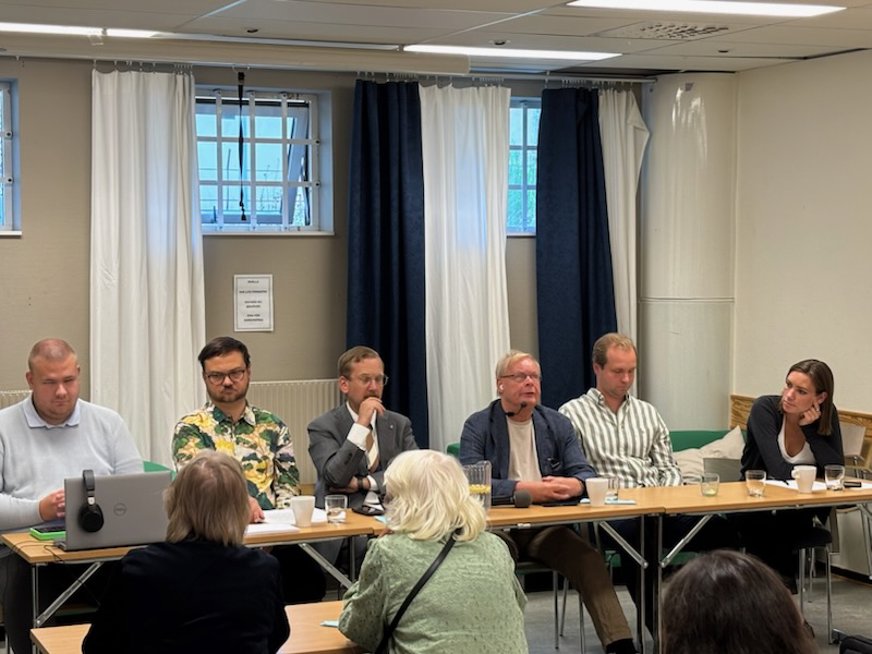 Valdebatt 19 aug. 6 Personer sitter vid ett långbord längst fram vända mot åhörare, i bakgrunden finns fönster med gardiner. Personerna vid bordet är Alexander Ojanne från Socialdemokraterna, Gabriel Kroon från Sverigedemokraterna, Nike Örbrink från Kristdemokraterna, Sverre Dahlstedt Eriksson från Miljöpartiet samt Kevin Kjelldahl och Urban Fernquist från SRF Stockholm Stad