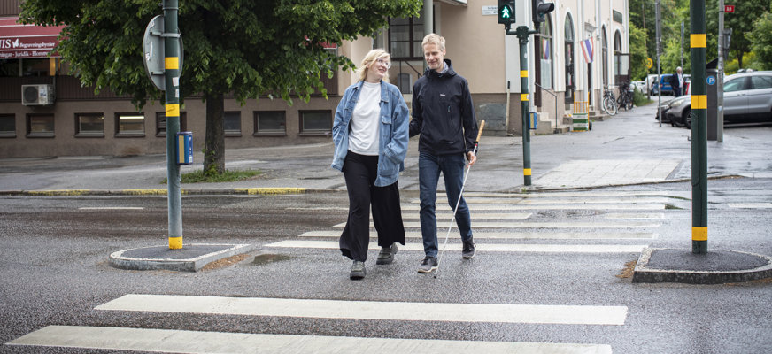 En kvinna och en man som går över ett övergångsställe. Kvinnan ledsagar mannen, och de skrattar tillsammans.