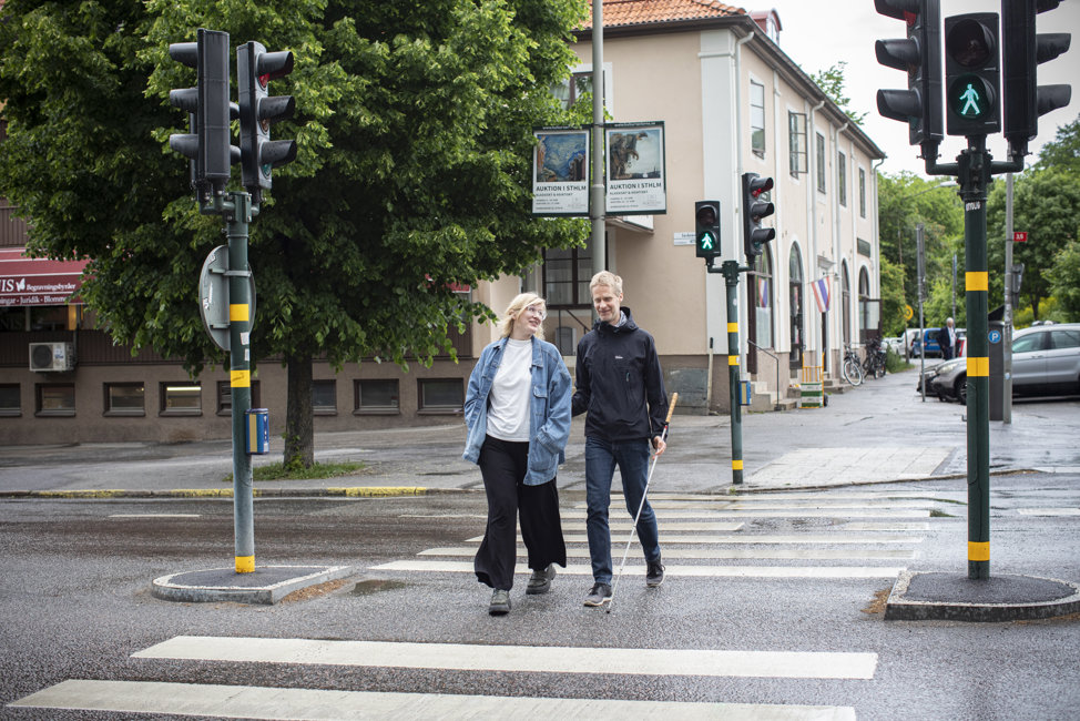 Man med käpp ledsagas av kvinna. De går över ett övergångsställe.
