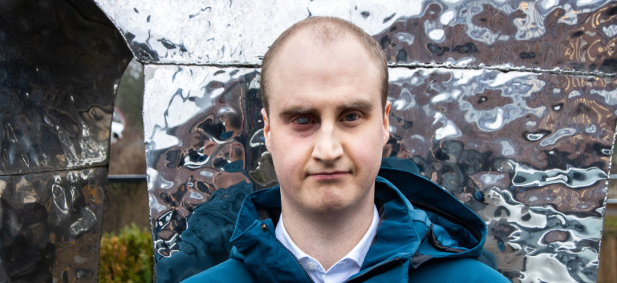 Närbild på Linus Johansson, en man med snaggat hår i 25-årsåldern. Linus står utomhus, framför en metallisk skulptur. Han har ett sammanbitet ansiktsuttryck och fäster blicken i kameran.