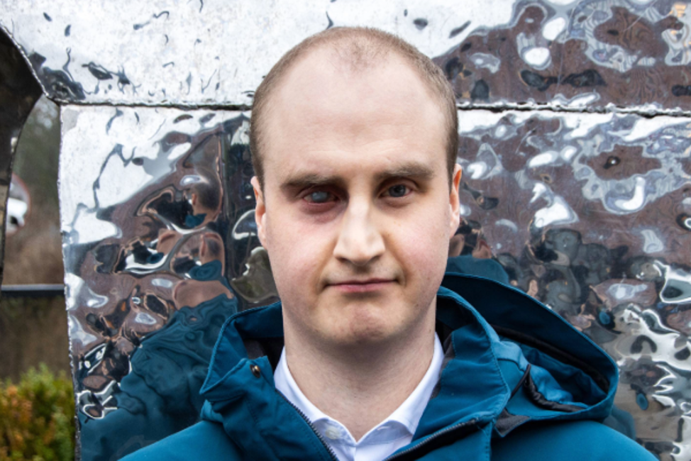 Närbild på Linus Johansson, en man med snaggat hår i 25-årsåldern. Linus står utomhus, framför en metallisk skulptur. Han har ett sammanbitet ansiktsuttryck och fäster blicken i kameran.