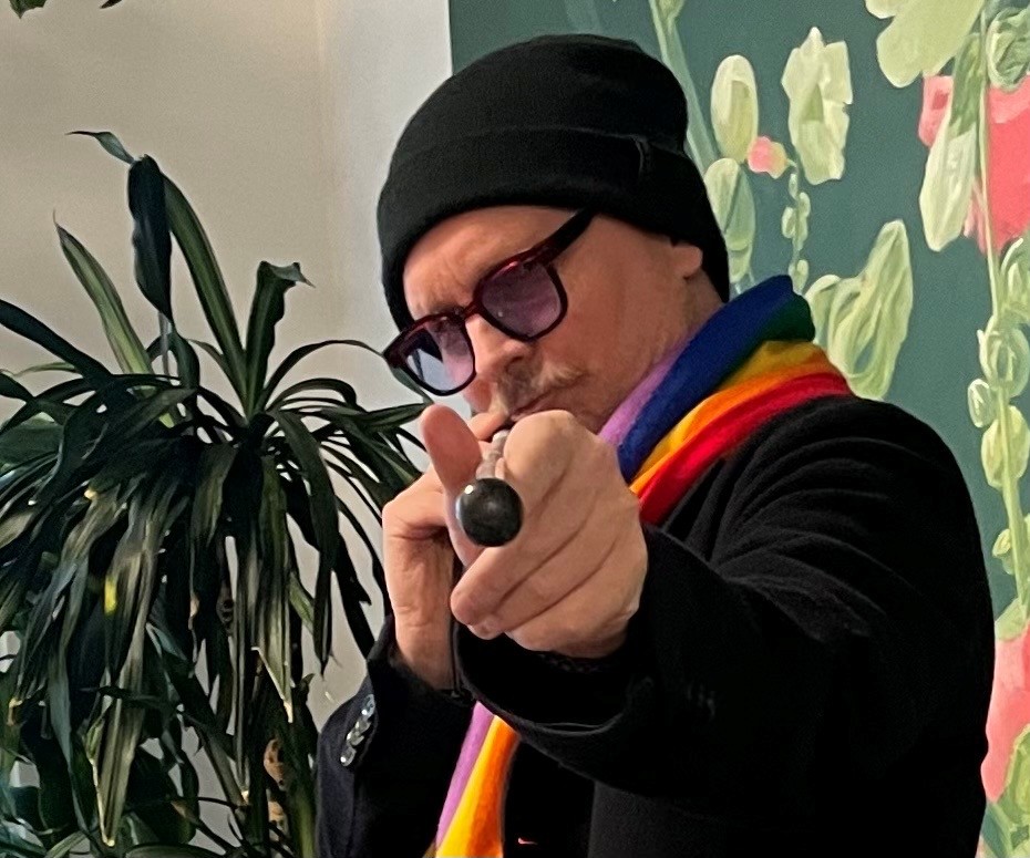 Mathias pekar mot kameran med sin vita käpp, som om han siktar med en biljardkö. Han är en medelålders man med mustasch som bär svarta glasögon med kraftiga bågar och tonade glas. Han har på sig en svart mössa och tröja, och bär en regnbågsrandig halsduk.