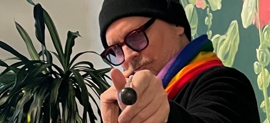 Mathias pekar mot kameran med sin vita käpp, som om han siktar med en biljardkö. Han är en medelålders man med mustasch som bär svarta glasögon med kraftiga bågar och tonade glas. Han har på sig en svart mössa och tröja, och bär en regnbågsrandig halsduk.