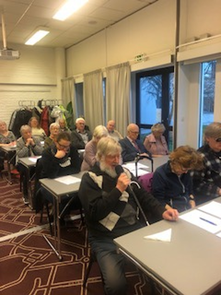 Bo Östen Svensson ordförande för SRF Uppsala-Knivsta sitter vid ett bord och pratar i en mikrofon. I bakgrunden syns andra mötesdeltagare vid olika bord.