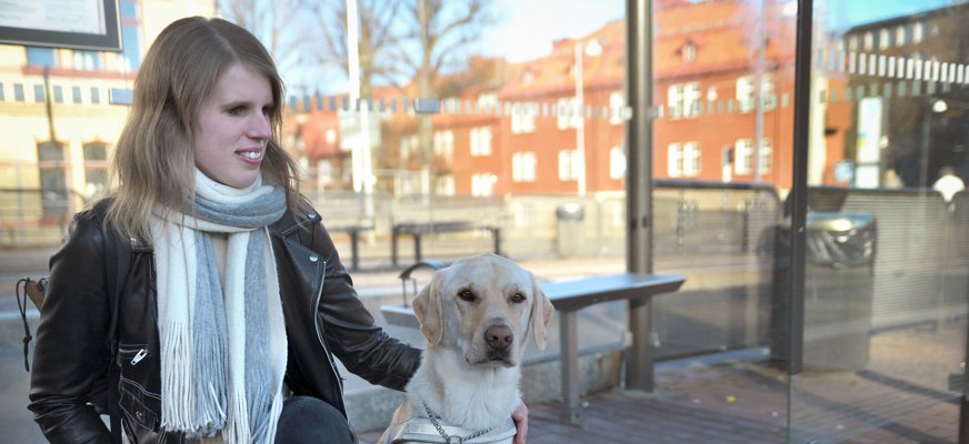 Viktoria sitter på knä bredvid sin ledarhund Poppe som är en ljus jaktlabrador. Viktora är i 25-årsålden och har skinnjacka och halsduk, hon har blicken riktad mot Poppe och ser glad ut.