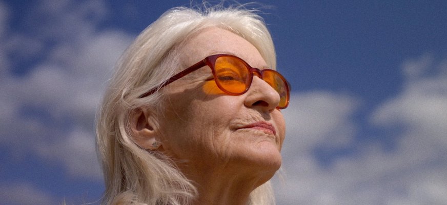 Alexandra Charles har vitt, axellångt hår och bär tonade solglasögon. Hon har ansiktet vänt mot solen och blundar. I bakgrunden syns en blå himmel med vita moln.