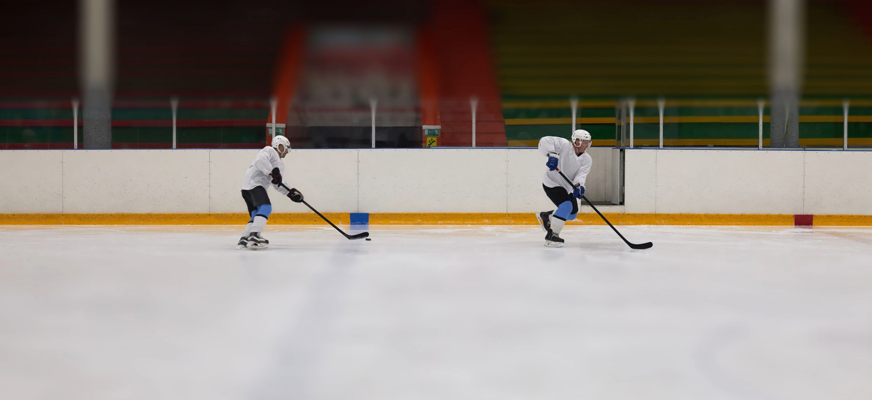 Två ishockeyspelare i vita tröjor, svarta byxor och vita hjälmar åker över isen med varsin klubba. Spelaren till höger har blå handskar och ligger längre fram, medan spelaren till vänster följer efter. En svart puck ligger på isen mellan dem. I bakgrunden syns sargen och läktare med röda, gröna och gula färgfält.