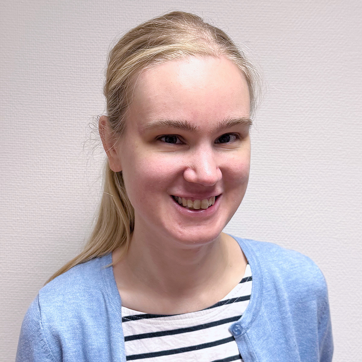 Porträttbild på Tova. Hon har långt blont hår med sidbena uppsatt i en hästsvans i nacken, markerade ögonbryn och blå ögon. Tova är klädd i en vit- och svartrandig tröja med blå kofta över. Hon ler och tittar rakt in i kameran. 