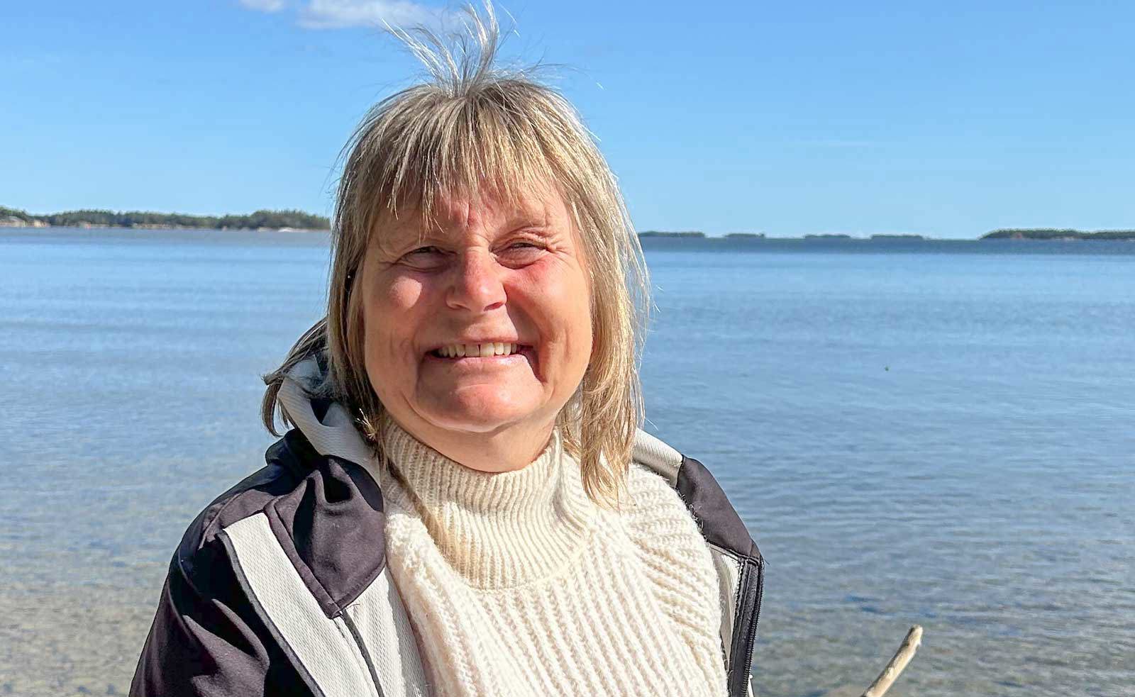 Medelålders kvinna står i solen med ett  glittrande vattendrag som bakgrund. Kvinnan har axellångt ljust hår med lugg och är klädd i vit polotröja med mörk vindjacka över. Hon ler mot kameran.