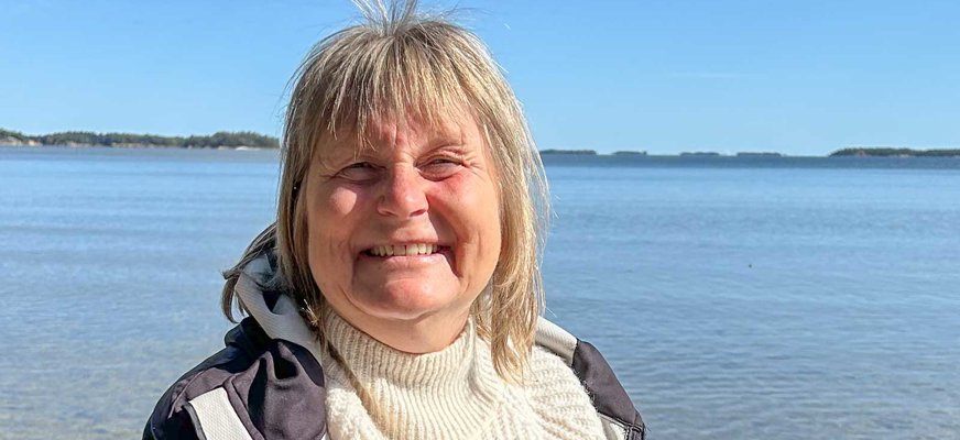 Medelålders kvinna står i solen med ett glittrande vattendrag som bakgrund. Kvinnan har axellångt ljust hår med lugg och är klädd i vit polotröja med mörk vindjacka över. Hon ler mot kameran.