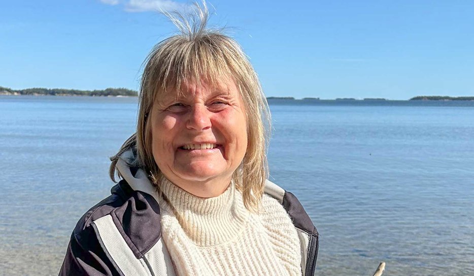 Medelålders kvinna står i solen med ett glittrande vattendrag som bakgrund. Kvinnan har axellångt ljust hår med lugg och är klädd i vit polotröja med mörk vindjacka över. Hon ler mot kameran.