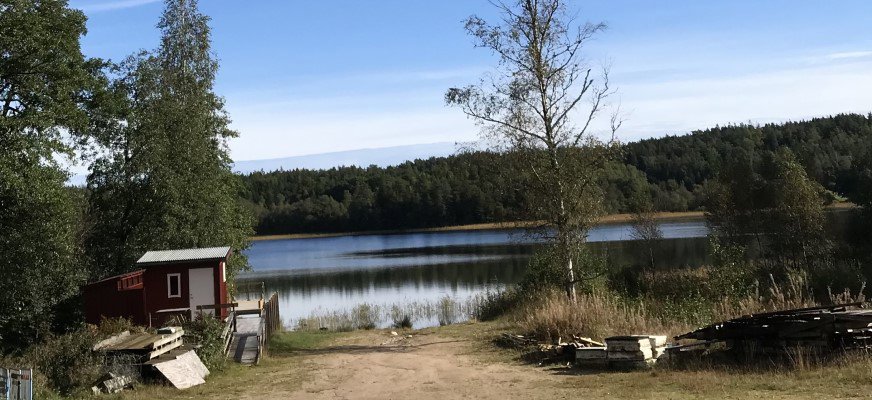 Grinnerödsjön är spegelblank och omgiven av skogsklädda kullar. Strandlinjen kantas av vass och sjöns lugna yta reflekterar den klarblå himlen. Till vänster finns en liten röd stuga med vitt tak och vit dörr, omgiven av träd och buskar.
