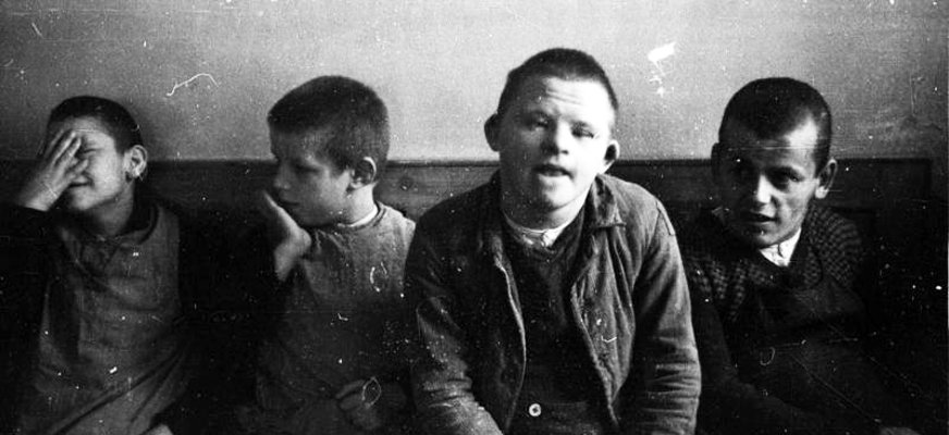 En äldre, svartvit bild föreställande fyra pojkar i 40-talskläder som sitter på en bänk tillsammans. En av dem håller en hand för sina ögon, en annan har tydliga ansiktsdrag av Downs syndrom.