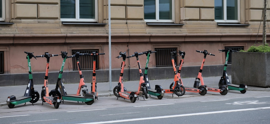 Flera elsparkcyklar står uppställda i rad längs en trottoar utanför en byggnad med stora fönster.