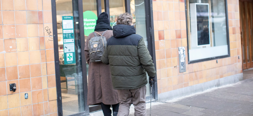 Två personer är på väg in genom entrén till ett apotek. Den ena bär ryggsäck och lång kappa, den andra en vinterjacka och använder käpp.