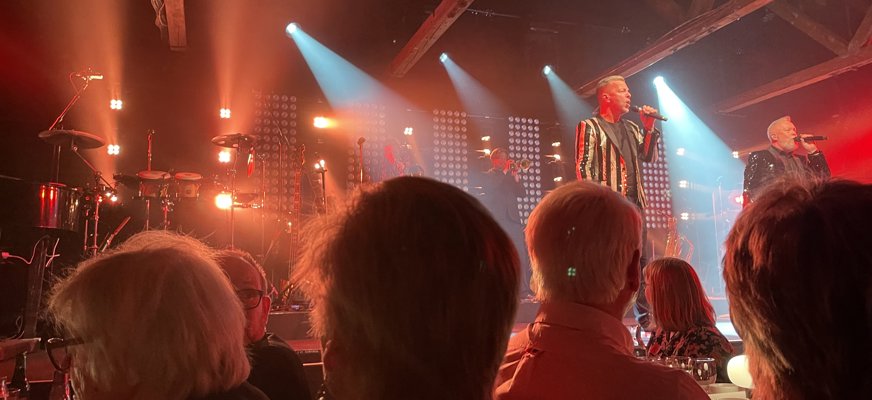 Bilden visar en spelning med Arvingarna. I förgrunden syns några människor som tittar mot scenen där Arvingarna står. Ljuset är rött