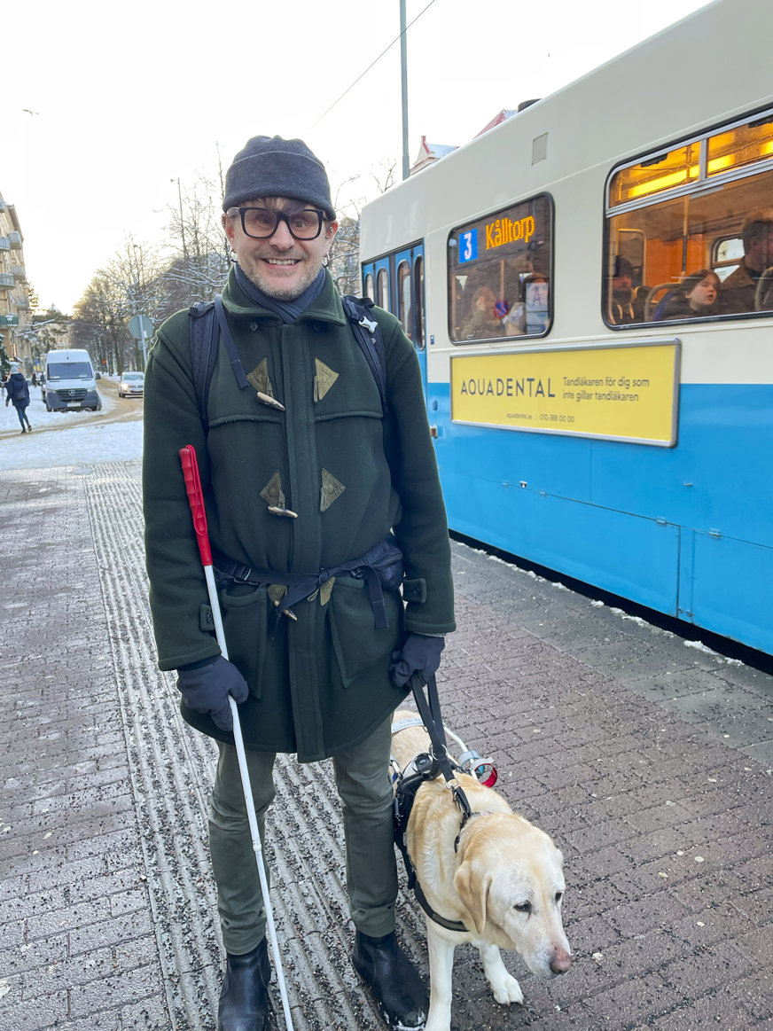 Finn står i en gatumiljö med sin gula labrador iförd sele. Han är klädd i blå jacka och mössa samt jeans och håller i en vit käpp. Bakom honom syns en blå spårvagn. 